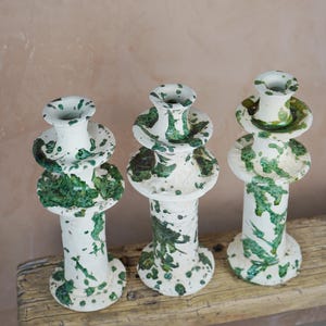 May include: Three ceramic candle holders with a white base and green splatter design. The holders have a tiered design with a wide base, a central column, and a top cup. They are displayed on a weathered wooden surface.