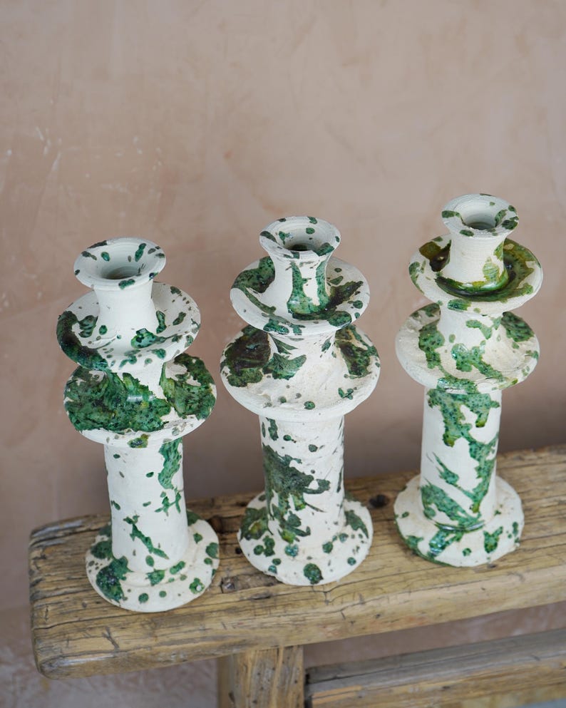 May include: Three ceramic candle holders with a white base and green splatter design. The holders have a tiered design with a wide base, a central column, and a top cup. They are displayed on a weathered wooden surface.