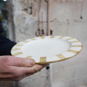 Peut inclure: Une assiette en céramique blanche avec une bordure à rayures beiges. L'assiette est tenue dans la main d'une personne, mettant en valeur son design. L'assiette fait partie d'une collection de vaisselle.