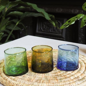 May include: Three glass tumblers in different colours: green, brown, and blue. The tumblers are sitting on a woven straw placemat.