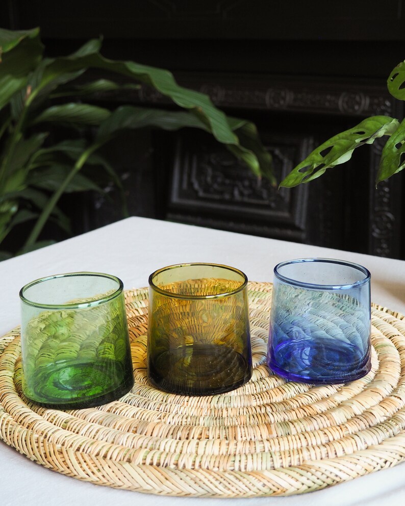 May include: Three glass tumblers in different colours: green, brown, and blue. The tumblers are sitting on a woven straw placemat.