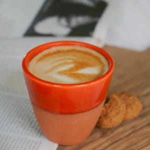 May include: A two-toned ceramic cup featuring a glossy orange upper section and a matte terracotta base, filled with a coffee beverage topped with white foam art. Two small, round, textured brown cookies sit beside the cup.