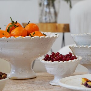May include: A collection of white ceramic pedestal bowls with scalloped edges. The largest bowl is filled with bright orange tangerines, while a smaller bowl contains a cluster of fresh red currants. The bowls feature a textured, rustic finish.