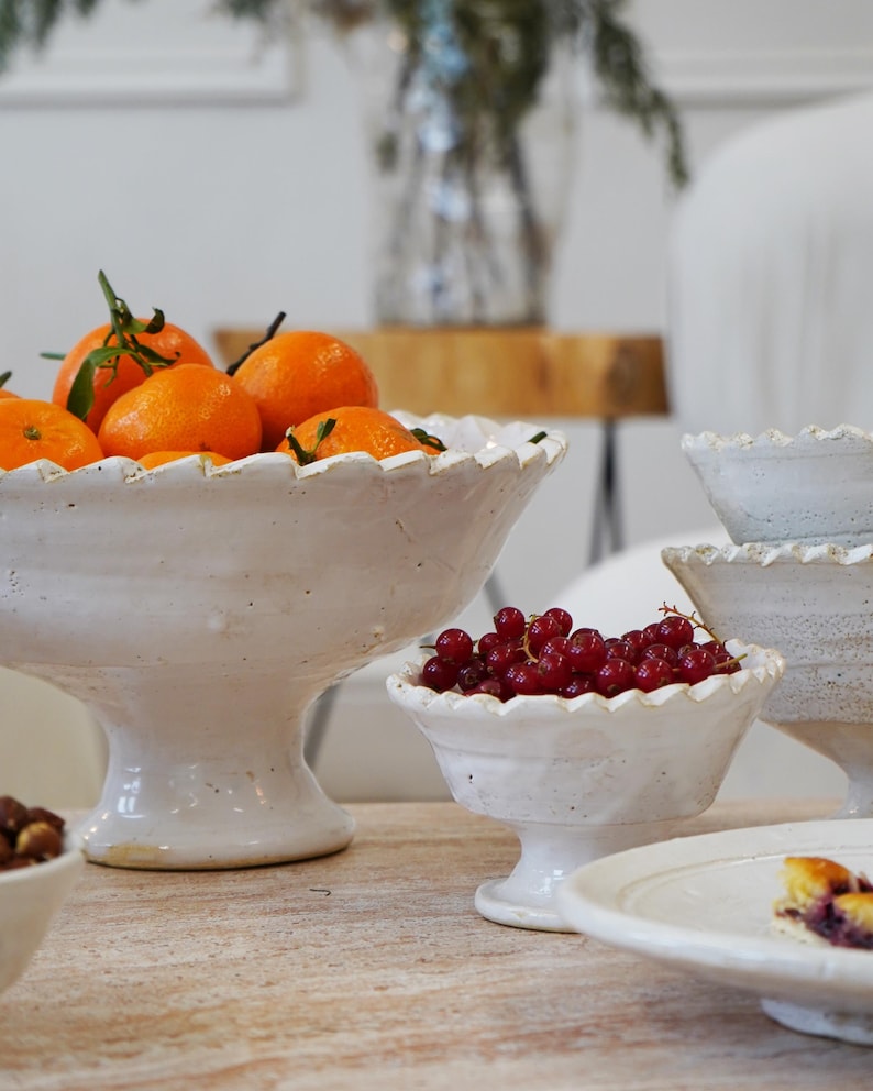 May include: A collection of white ceramic pedestal bowls with scalloped edges. The largest bowl is filled with bright orange tangerines, while a smaller bowl contains a cluster of fresh red currants. The bowls feature a textured, rustic finish.