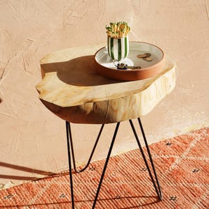 Pode incluir: Uma mesa lateral de madeira natural com pernas de metal preto em forma de grampo. Um pequeno recipiente de cerâmica com listras verdes e brancas contém fósforos. Uma bandeja branca e marrom contém chaves e moedas. A mesa está sobre um tapete laranja estampado.