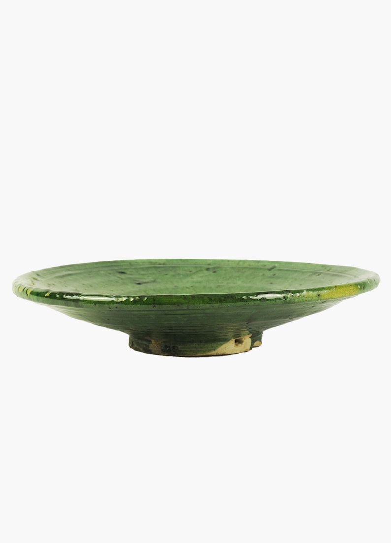 May include: A green ceramic bowl with a wide, shallow design. The bowl has a slightly raised base and a glossy finish. The color is a vibrant, solid green, with some variations in tone. The bowl's diameter is approximately 12 inches.