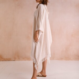 Puede incluir: Un poncho de algod&oacute;n blanco con detalles de flecos. El poncho tiene capucha y lo lleva puesto una persona de pie sobre un suelo blanco.