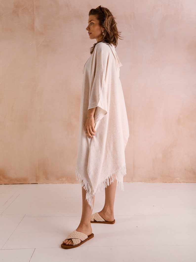 Puede incluir: Un poncho de algod&oacute;n blanco con detalles de flecos. El poncho tiene capucha y lo lleva puesto una persona de pie sobre un suelo blanco.