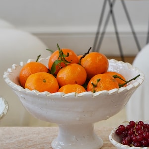 May include: A white ceramic pedestal bowl with a scalloped rim, filled with bright orange tangerines that have green leaves attached. The bowl has a textured, rustic finish and sits on a short, sturdy base, presenting a classic fruit display.