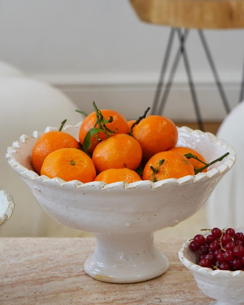 May include: A white ceramic pedestal bowl with a scalloped rim, filled with bright orange tangerines that have green leaves attached. The bowl has a textured, rustic finish and sits on a short, sturdy base, presenting a classic fruit display.