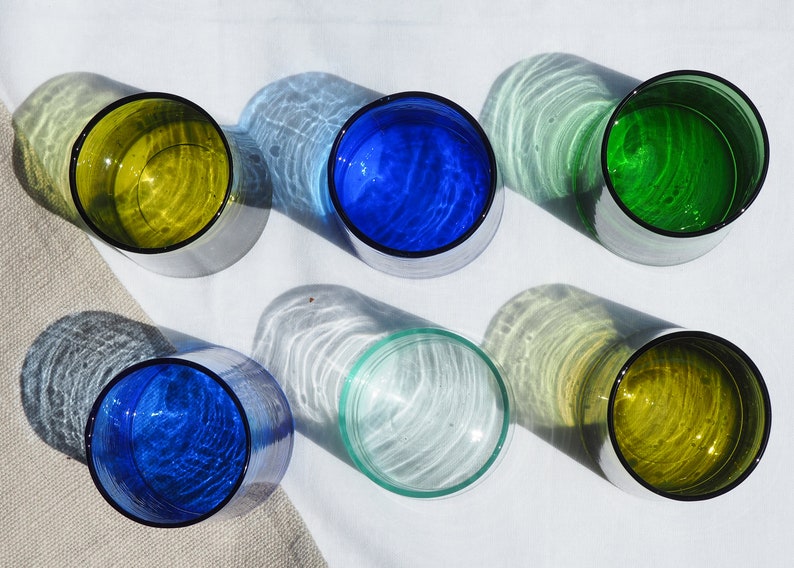 Peut inclure: Six verres en verre de diff&eacute;rentes couleurs, dont bleu, vert et jaune. Les verres sont dispos&eacute;s en cercle sur une surface blanche.