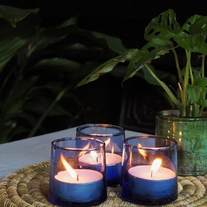 May include: Three blue glass candle holders with white tea lights burning inside. The candle holders are on a woven wicker placemat. A green potted plant is in the background.