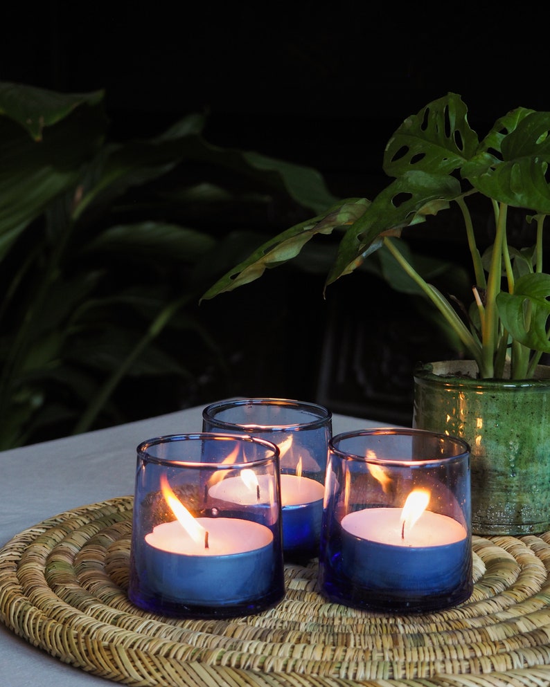 May include: Three blue glass candle holders with white tea lights burning inside. The candle holders are on a woven wicker placemat. A green potted plant is in the background.