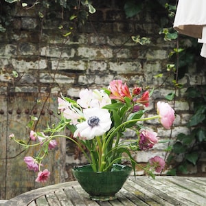 Op de afbeelding: Een groene keramische kom gevuld met een bloemstuk van roze, witte en rode bloemen. De bloemen zijn in een vaas op een houten tafel gerangschikt.