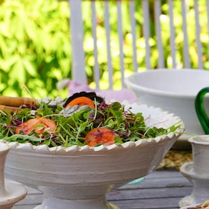 May include: A large, off-white ceramic pedestal bowl with a scalloped rim, filled with a fresh green salad featuring sliced red tomatoes and purple microgreens. The bowl has a textured, hand-crafted appearance and sits on a sturdy, wide base.