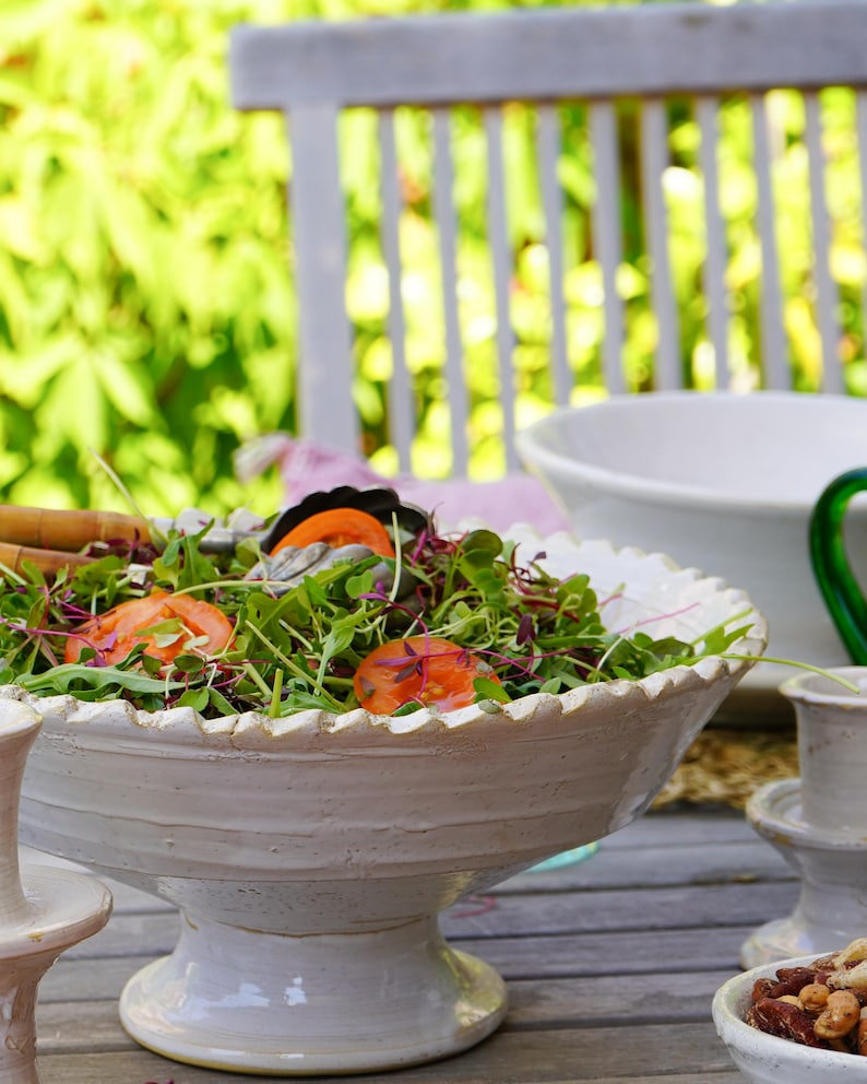 May include: A large, off-white ceramic pedestal bowl with a scalloped rim, filled with a fresh green salad featuring sliced red tomatoes and purple microgreens. The bowl has a textured, hand-crafted appearance and sits on a sturdy, wide base.