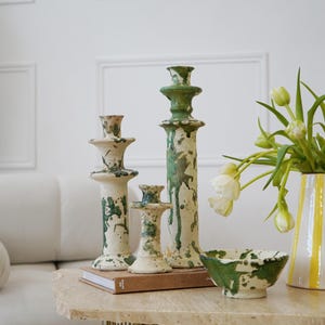 Op de afbeelding: Een verzameling keramische woondecoratie-items, waaronder drie kandelaars en een kleine kom, allemaal met een crèmekleurige basis en een groen spatterontwerp. Er is ook een vaas met gele strepen en witte tulpen.
