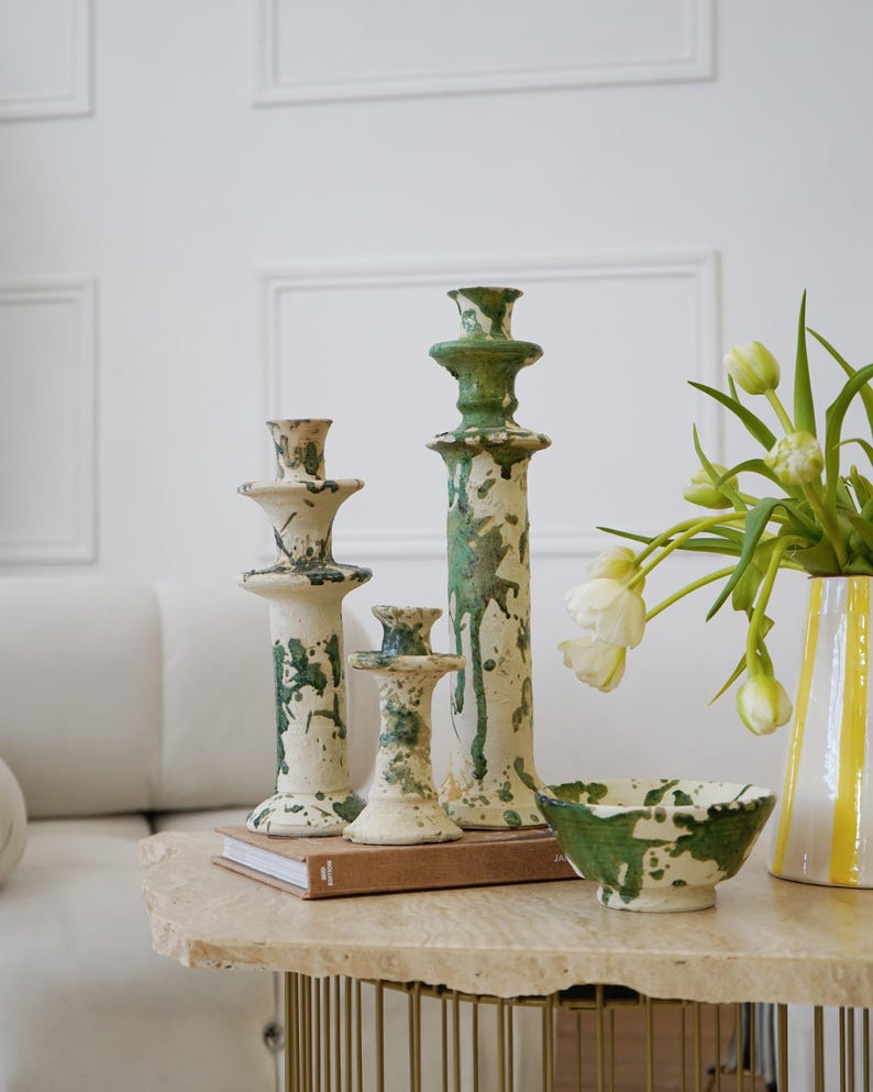 May include: A collection of ceramic home decor items, including three candlestick holders and a small bowl, all with a cream base and green splatter design. A vase with yellow stripes and white tulips is also present.