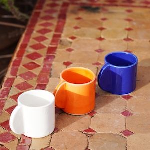 Pode incluir: Três canecas de cerâmica em cores sólidas branco, laranja brilhante e azul profundo. Cada caneca apresenta um corpo cilíndrico liso e uma pequena alça arredondada e saliente na lateral. As canecas estão dispostas em linha diagonal.