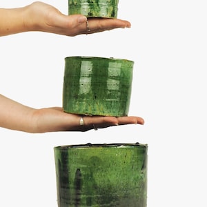 May include: Three green ceramic pots of varying sizes stacked on top of each other. The pots are all glazed in a dark green color with a slightly textured surface.