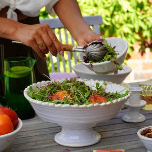May include: A white ceramic pedestal bowl with a scalloped rim filled with a salad of mixed greens and sliced tomatoes. The bowl features a textured, rustic finish and sits on a circular base. A metal serving utensil with a wooden handle is present.