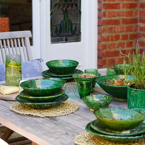 May include: A collection of emerald green glazed ceramic dinnerware including stacked bowls and plates, a large salad bowl, and small drinking glasses arranged on a wooden table with woven placemats and a glass pitcher of water.
