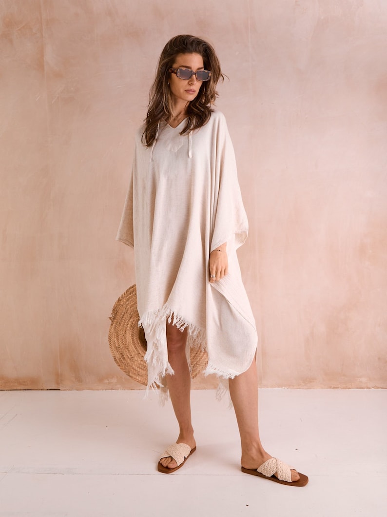 Puede incluir: Un poncho de algod&oacute;n blanco con detalles de flecos y capucha con cord&oacute;n. El poncho lo lleva puesto una persona que lleva gafas de sol y sandalias.