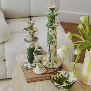 May include: A collection of ceramic home decor items, including three candle holders of varying heights and a small bowl. The pieces are off-white with green splatter paint. A book titled "The World Atlas of Coffee" is also visible.