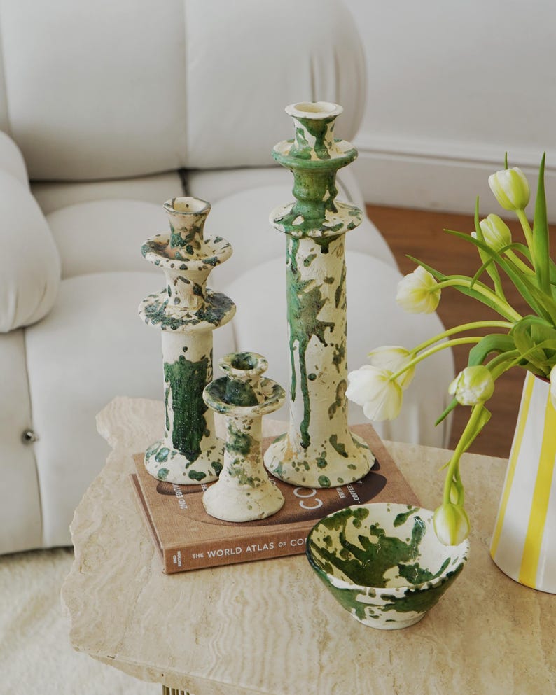 May include: A collection of ceramic home decor items, including three candle holders of varying heights and a small bowl. The pieces are off-white with green splatter paint. A book titled "The World Atlas of Coffee" is also visible.