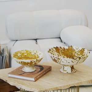 May include: Two decorative ceramic bowls with a white base and a yellow and brown splatter design. The bowls are on a light-colored stone table with a book. The table has a gold metal base.
