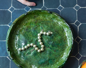 Op de afbeelding: Een zilveren armband met ronde kralen ligt op een groene keramische schaal. De schaal heeft een getextureerd oppervlak en staat op een blauwe betegelde achtergrond.