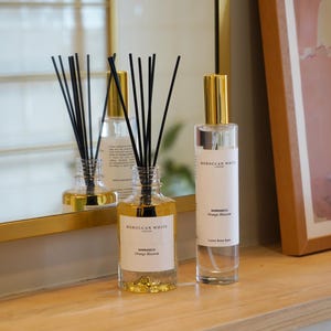 May include: A clear glass reed diffuser with black reeds and gold-toned decorative flakes at the base, alongside a tall clear glass room spray bottle with a gold cap. Both feature white labels reading Moroccan White London Marrakech Orange Blossom.