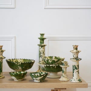 May include: A collection of ceramic bowls and candle holders with a green and white splatter design. The bowls have a ruffled edge. The candle holders are various heights and shapes. The items are displayed on a light wood surface.