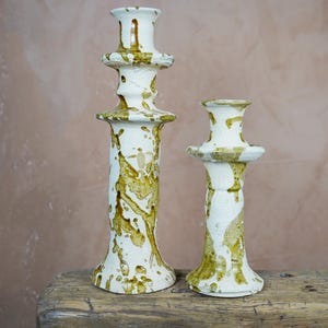 May include: Two ceramic candle holders with a white base and olive green splatter design. The candle holders are different sizes, with the taller one measuring approximately 12 inches in height. They sit on a rustic wooden surface.