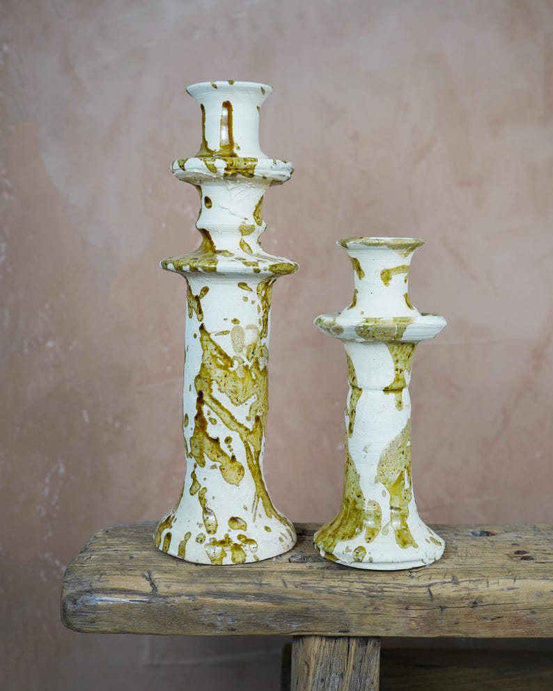 May include: Two ceramic candle holders with a white base and olive green splatter design. The candle holders are different sizes, with the taller one measuring approximately 12 inches in height. They sit on a rustic wooden surface.