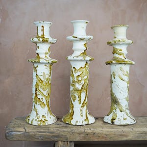 May include: Three white ceramic candle holders with a textured, dripped glaze in shades of brown and gold. The candle holders are displayed on a rustic wooden surface against a neutral background. The design is artistic and decorative.