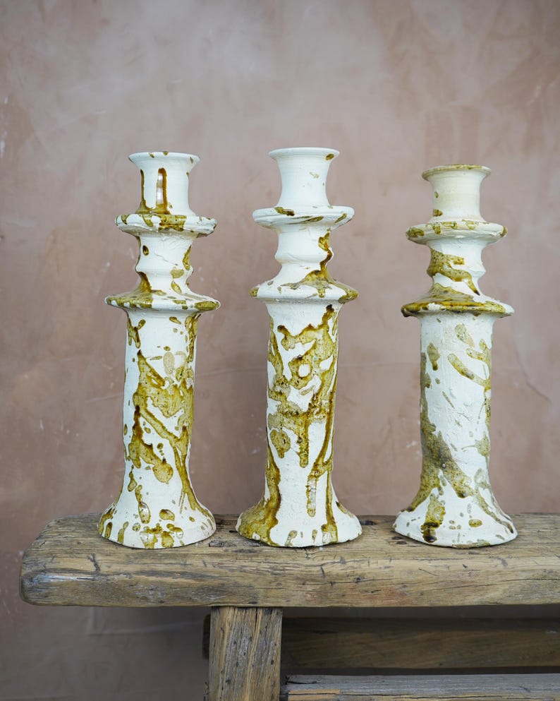 May include: Three white ceramic candle holders with a textured, dripped glaze in shades of brown and gold. The candle holders are displayed on a rustic wooden surface against a neutral background. The design is artistic and decorative.