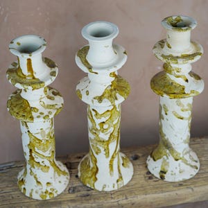 May include: Three white ceramic candle holders with a speckled olive green glaze. The candle holders have a tiered design with a wide base and a narrow top. They are displayed on a weathered wooden surface.