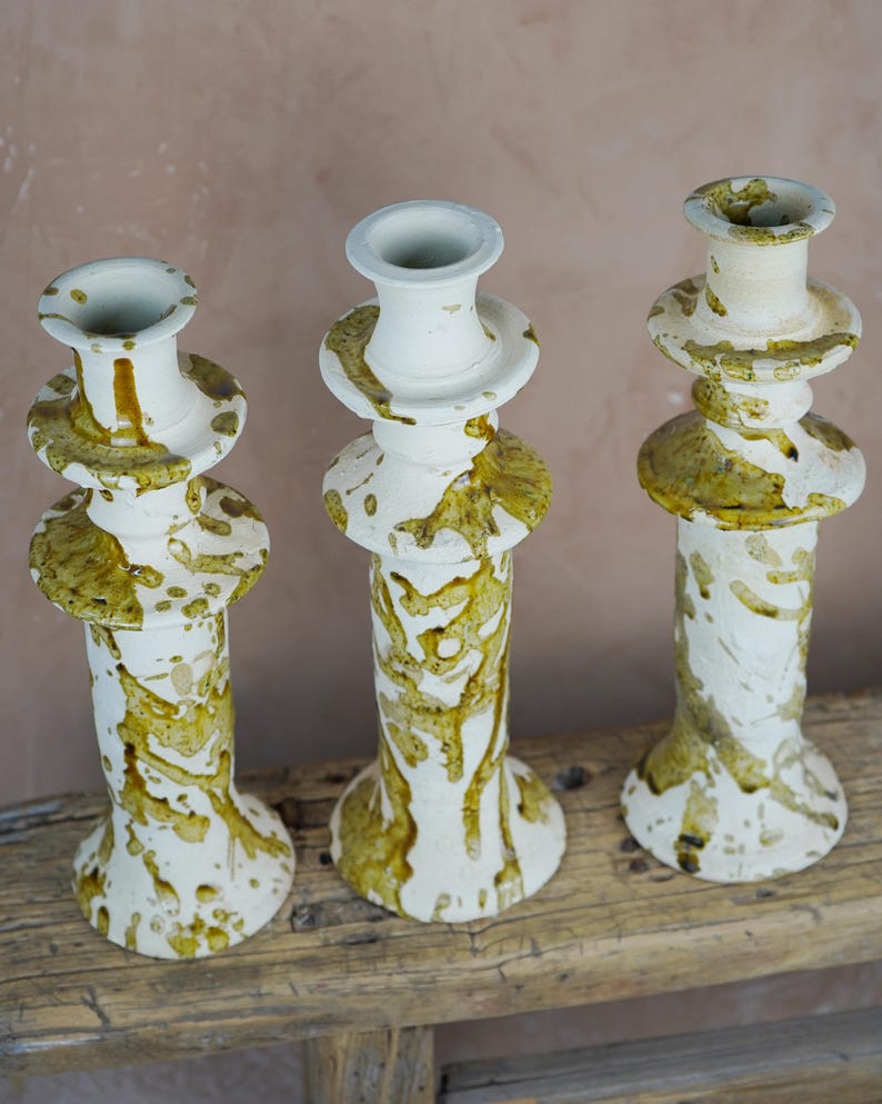 May include: Three white ceramic candle holders with a speckled olive green glaze. The candle holders have a tiered design with a wide base and a narrow top. They are displayed on a weathered wooden surface.