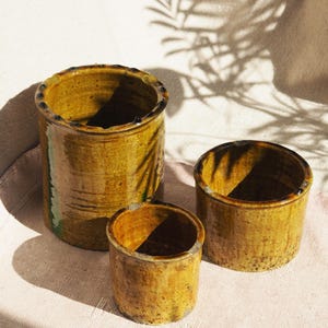 May include: Three ceramic pots in various sizes, all with a yellow-brown glaze and a rough, textured surface. The pots are arranged on a white fabric background.