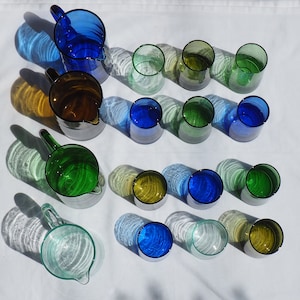 Peut inclure: Une collection de pichets et de verres en verre dans diff&eacute;rentes nuances de bleu, de vert et de transparent. Les pichets ont une forme arrondie et une poign&eacute;e, tandis que les verres sont cylindriques. Les verres sont dispos&eacute;s sur une surface blanche.