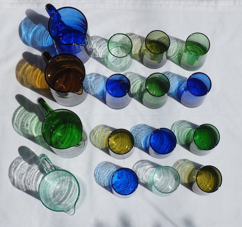 Peut inclure: Une collection de pichets et de verres en verre dans diff&eacute;rentes nuances de bleu, de vert et de transparent. Les pichets ont une forme arrondie et une poign&eacute;e, tandis que les verres sont cylindriques. Les verres sont dispos&eacute;s sur une surface blanche.