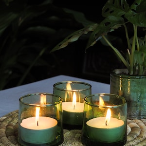 May include: Four green glass candle holders with white tea lights burning inside. The candle holders are on a woven straw placemat.