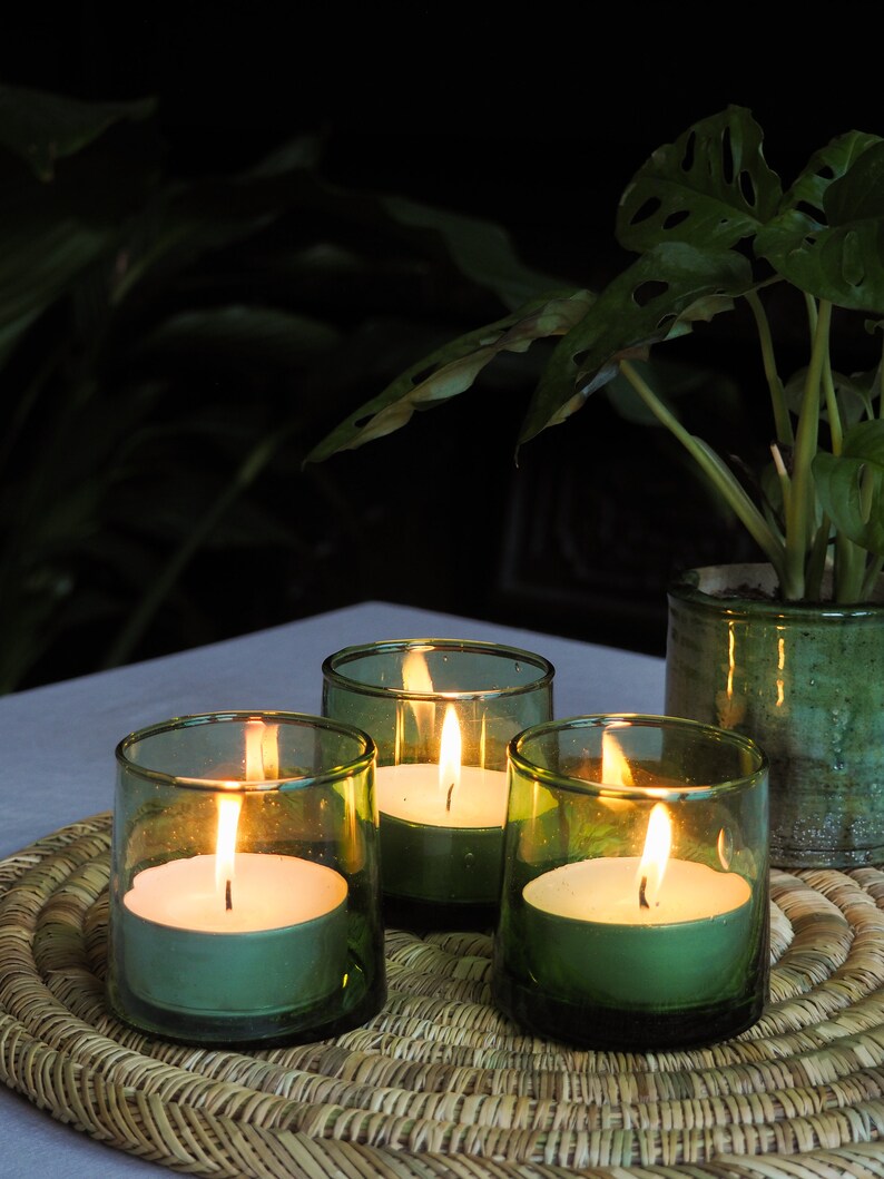 May include: Four green glass candle holders with white tea lights burning inside. The candle holders are on a woven straw placemat.