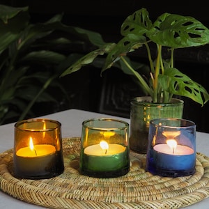 May include: Three glass candle holders with lit candles. The candle holders are brown, green, and blue. The candles are white and are sitting on a woven wicker mat.