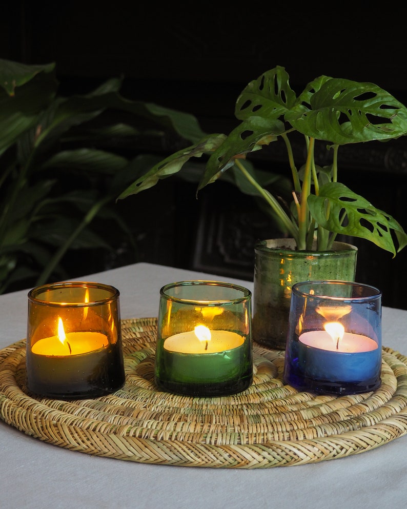 May include: Three glass candle holders with lit candles. The candle holders are brown, green, and blue. The candles are white and are sitting on a woven wicker mat.