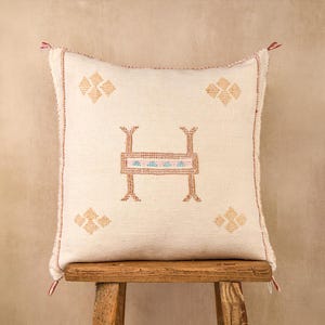 May include: Cream-colored square pillow with an embroidered design. The pillow features a central geometric design in brown, red, and blue, surrounded by four smaller floral-like patterns. Red tassels adorn the corners.