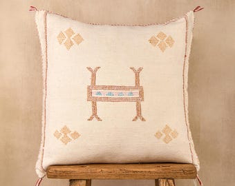 May include: Cream-colored square pillow with an embroidered design. The pillow features a central geometric design in brown, red, and blue, surrounded by four smaller floral-like patterns. Red tassels adorn the corners.