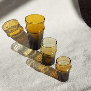 May include: Three amber glass tumblers of varying sizes, with a textured surface, sitting on a white linen tablecloth.