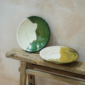 Puede incluir: Dos platos de cer&aacute;mica hechos a mano con una est&eacute;tica r&uacute;stica. Uno es redondo con un esmalte verde y blanco, el otro es ovalado con un esmalte amarillo y blanco. Descansan sobre un banco de madera envejecida contra una pared neutra.